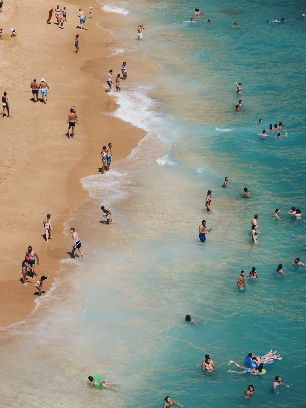 People enjoying a beach and pool area with clear blue water and sandy shore.