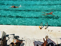 Two people swimming in a clear blue pool with lounge chairs and towels on the edge.