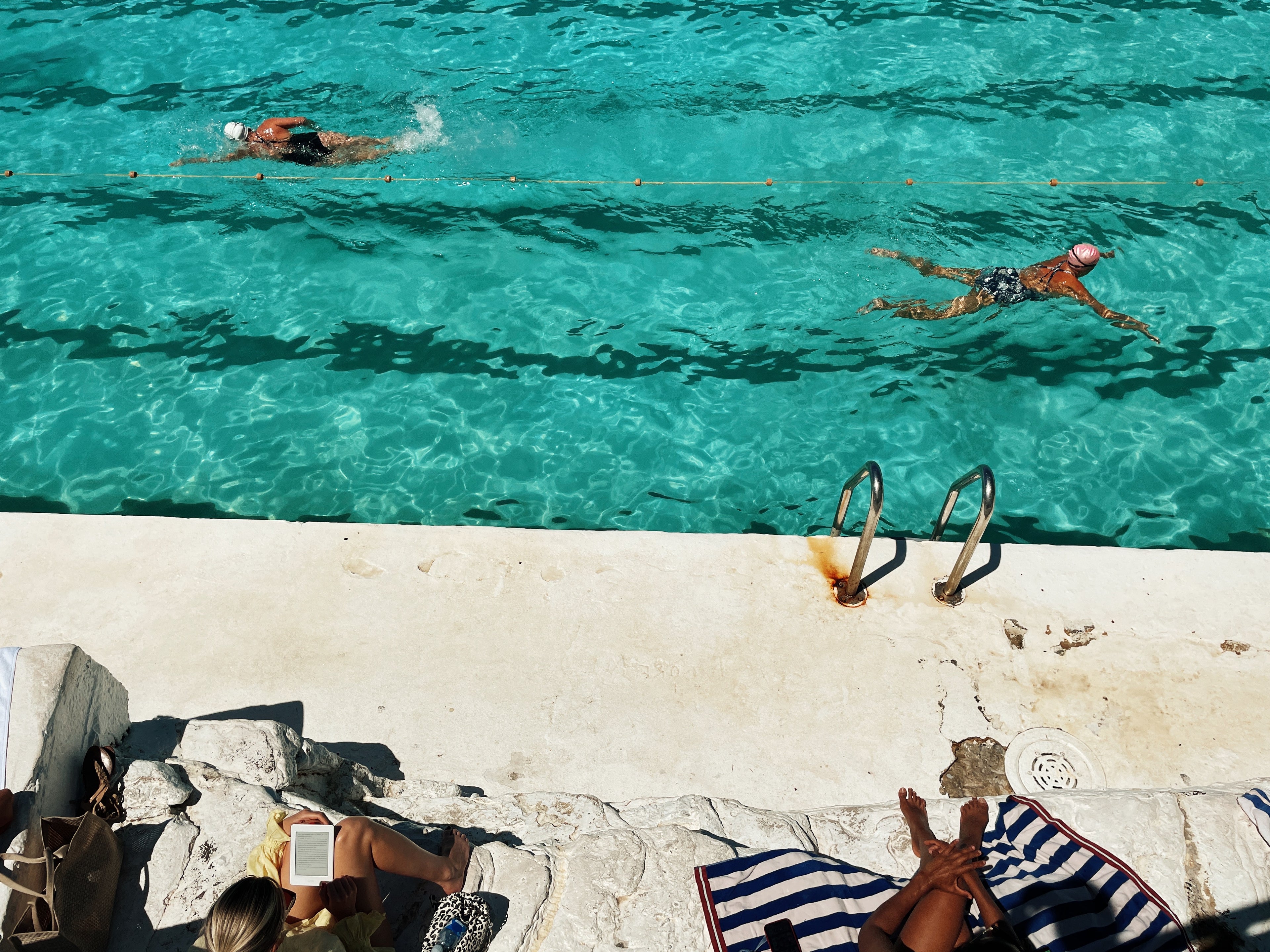 Two people swimming in a clear blue pool with lounge chairs and towels on the edge.