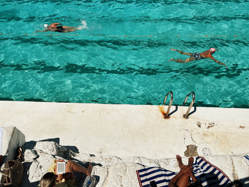 Two people swimming in a clear blue pool with lounge chairs and towels on the edge.