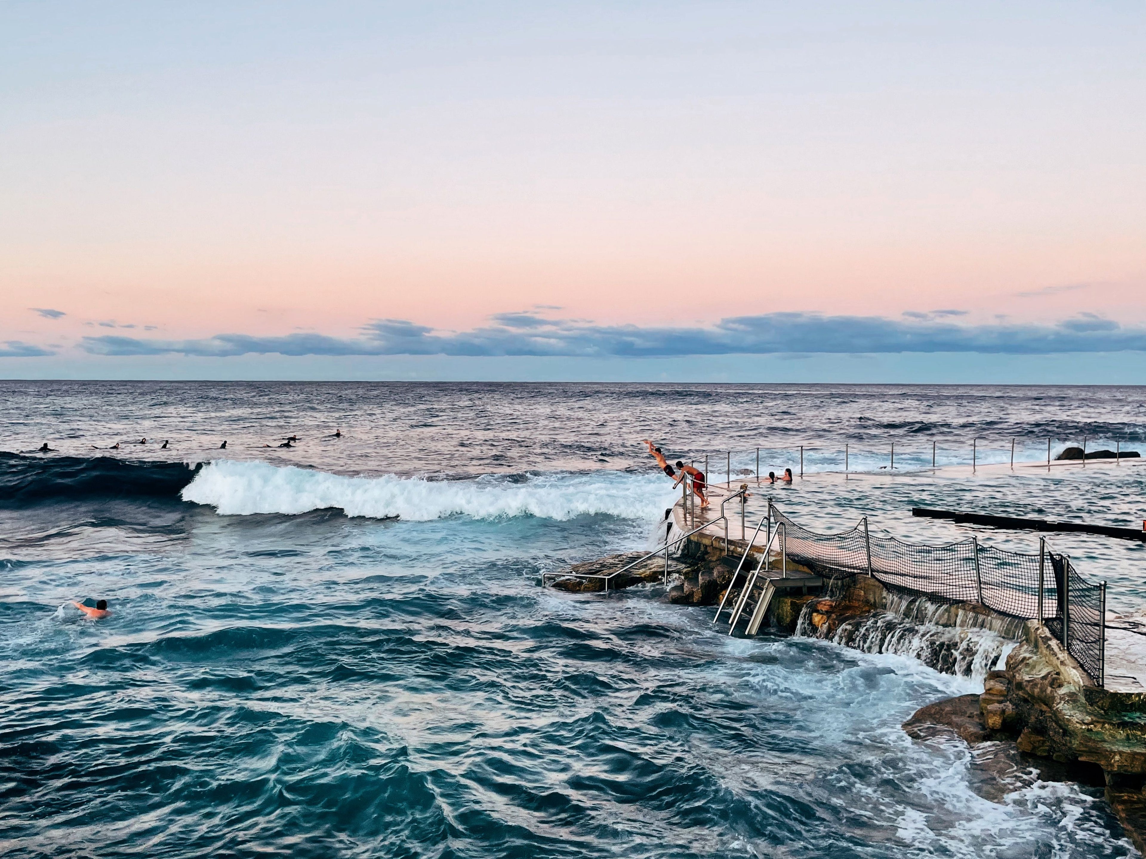 People enjoying a swim at a coastal pool with ocean waves and a dock.