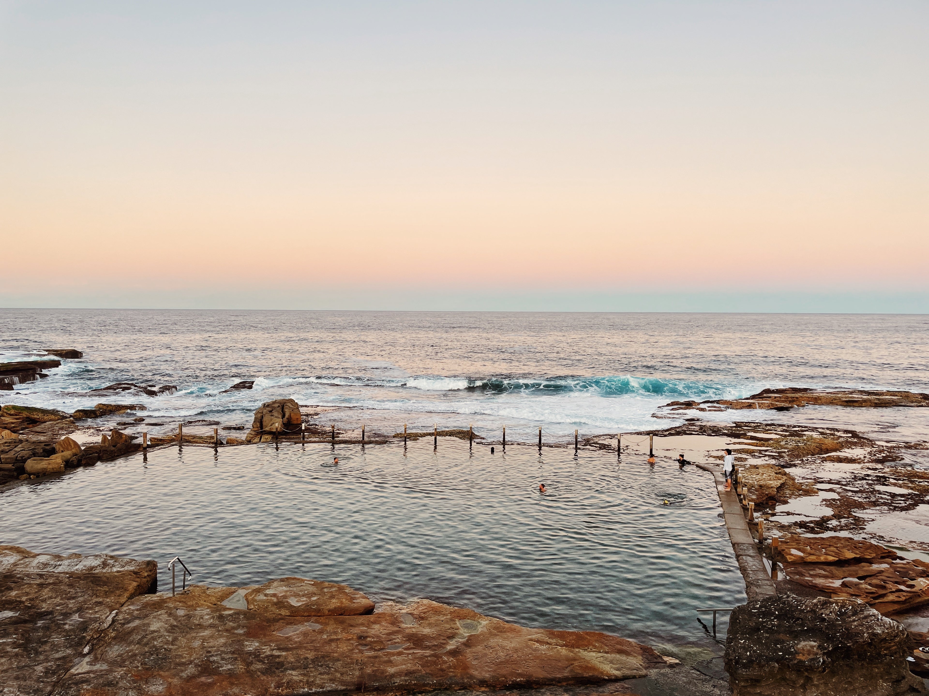 Natural rock pool by the ocean with people swimming at sunset.
