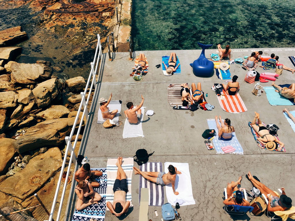 People sunbathing on towels by a poolside with a rocky cliff and greenery in the background