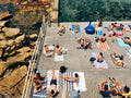 People sunbathing on towels by a poolside with a rocky cliff and greenery in the background