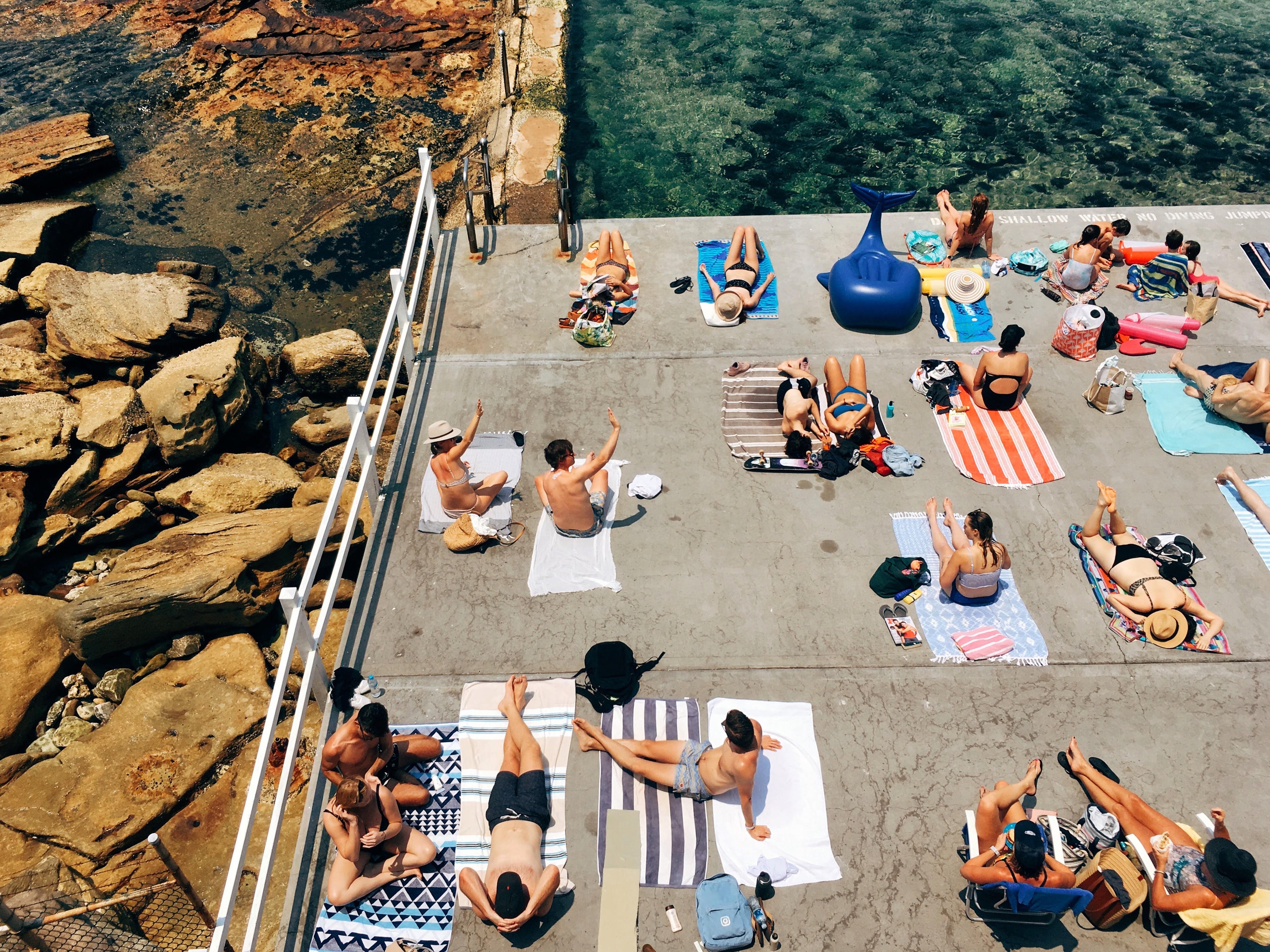 People sunbathing on towels by a poolside with a rocky cliff and greenery in the background
